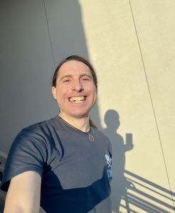 Christopher Nawoichik standing on stairs in sunset smiling wearing blue t-shirt with hair in a ponytail.
